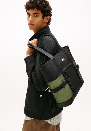 Junger Mann mit schwarzer Jacke, der eine schwarze und grüne Tragetasche mit dem Tommy Jeans-Logo über der Schulter trägt.