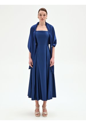 Femme debout vêtue d'une robe bleue longue sans bretelles avec un châle assorti et des sandales à talons hauts dorées, sur un fond blanc.