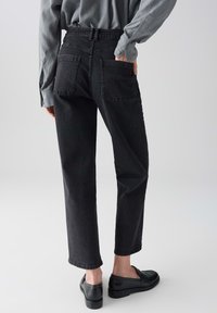 Persona che indossa jeans a vita alta neri e mocassini neri, con una mano nella tasca posteriore, su uno sfondo grigio uniforme.