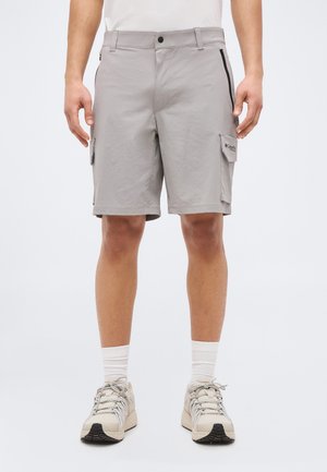 Mann trägt hellgraue Cargo-Shorts mit Seitentaschen und beigefarbene Sneakers mit weißen Socken, steht vor einem schlichten weißen Hintergrund.