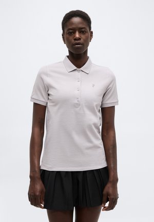 Femeie de culoare purtând un tricou polo gri deschis cu mâneci scurte, nasturi și un mic logo, asortat cu o fustă neagră plisată, stând în picioare pe fundal alb.