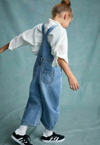 Lichtblauwe denim jumpsuit met een losse pasvorm, verstelbare schouderbanden en twee achterzakken. Gekoppeld met een witte blouse en zwarte sneakers.