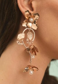 Orecchini in oro rosa con un design floreale caratterizzato da petali testurizzati, accenti di cristallo e elementi pendenti in perla. Forma curva e delicata.
