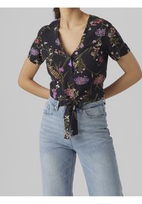 Blusa floral de manga corta, fondo negro, colores vibrantes, cierre de botones y detalle de lazo en la cintura, combinada con pantalones vaqueros azul claros.