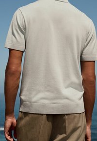 Lichtgrijze polo met korte mouwen, voorzien van een geribbelde zoom en kraag. Zachte textuur, fitted ontwerp. Zichtbare armen en een achtergrond van oceaan en lucht.
