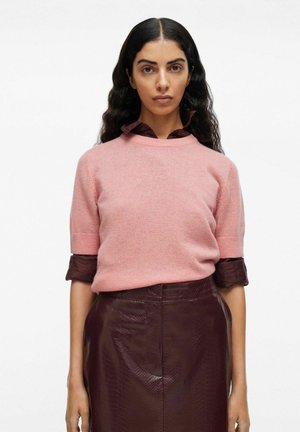Femme aux longs cheveux noirs ondulés portant un pull rose à manches courtes par-dessus une chemise boutonnée bordeaux et une jupe bordeaux texturée, se tenant contre un fond blanc.