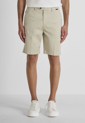Beige knielange shorts gedragen door een persoon die staat, gecombineerd met witte sneakers en een wit shirt, op een effen lichte achtergrond.