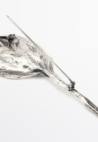 Silberfarbener Metallanhänger mit strukturiertem Oberflächen und Steckverschluss, der eine unregelmäßige, organische Form auf einem weißen Hintergrund zeigt.
