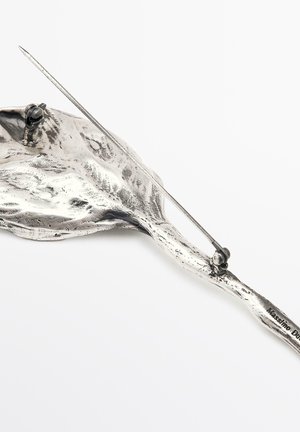 Spilla in metallo argento con superficie testurizzata e chiusura a spillo, caratterizzata da una forma irregolare e organica su uno sfondo bianco.
