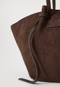 Braune Suede-Tote-Bag mit weicher Textur, konischer Form und schlanken Ledergriffen. Enthält ein sicheres, geknotetes Verschlussdetail.