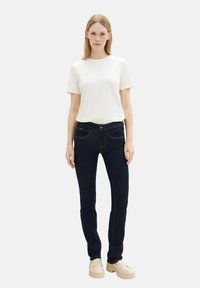 Jeans de mezclilla oscura con corte ajustado, que cuentan con cinco bolsillos y costuras en contraste, combinados con una camiseta blanca lisa. Zapatos gruesos en color beige.