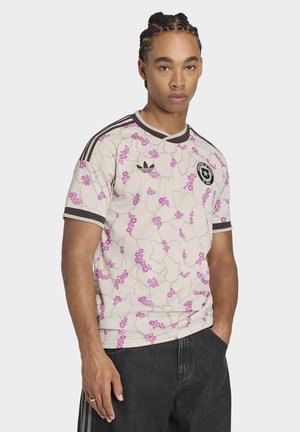 Jeune homme aux cheveux tressés portant un t-shirt Adidas beige à motifs floraux avec des bandes noires et un jean foncé, debout avec une main dans la poche.