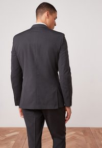 Veste de costume grise à texture lisse, fente unique à l'arrière et poignets à un bouton. Assortie à un pantalon coordonné sur un sol en bois.