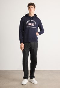 Granatowa bluza z kapturem z logo "GANT" w kolorze czerwonym i białym, w połączeniu z ciemnoniebieskimi dżinsami i białymi trampkami. Proste tło.