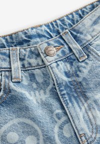 Shorts en denim bleu délavé avec une texture à motifs, dotés d'un bouton en métal avec "Authentic 1982" et des détails de couture visibles.