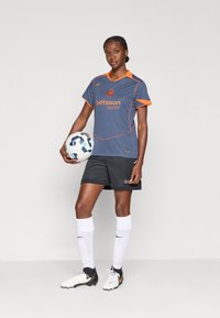 Grå fotbollströja med oranga detaljer och logotyp, svarta shorts, vita knästrumpor och vita fotbollsskor, hållande en fotboll.