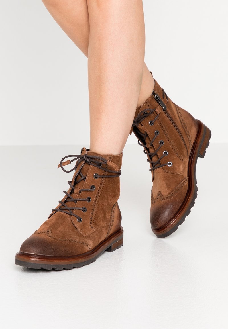 Bottines en daim marron à lacets avec détails brogue portées sur des jambes croisées sur un fond clair.