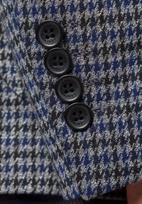 Tkanina w wzór houndstooth w odcieniach szaro-niebieskich, z czterema czarnymi guzikami z kwiatowym wzorem. Zbliżenie na szczegół kurtki.
