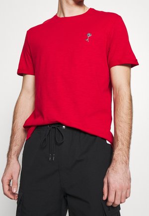 Homme portant un T-shirt rouge uni à manches courtes avec une petite broderie de rose noire et blanche sur la poitrine et un pantalon noir à cordon.