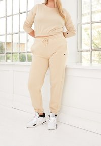 Frau trägt einen beigefarbenen, kurzen Pullover und passende Jogginghosen mit weißen High-Top-Sneakern, steht an einem großen Fenster in einem hellen Raum.