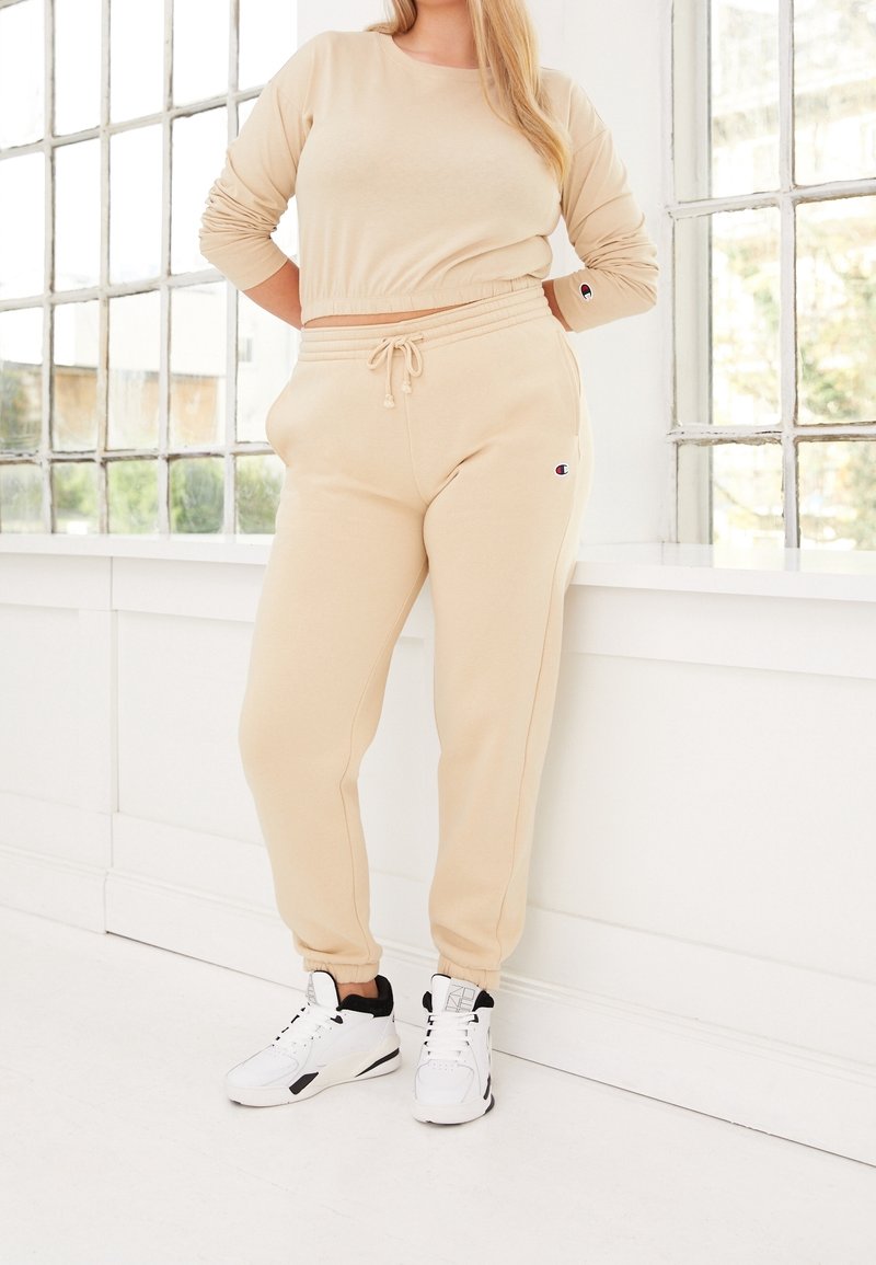 Frau trägt einen beigefarbenen, kurzen Pullover und passende Jogginghosen mit weißen High-Top-Sneakern, steht an einem großen Fenster in einem hellen Raum.