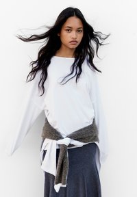 Weißes Langarmoberteil mit lockerer Passform, kombiniert mit einem grauen Strick-Wickelgurt. Grauer Rock reicht bis zu den Knöcheln. Der Hintergrund ist reinweiß.