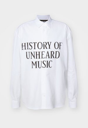 Vit långärmad skjorta med knappar som har "HISTORIA OM OHÖRD MUSIK" tryckt i fet, sliten svarts text på framsidan.