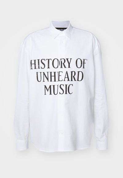Vit långärmad skjorta med knappar som har "HISTORIA OM OHÖRD MUSIK" tryckt i fet, sliten svarts text på framsidan.