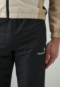 Pantaloni da jogging da uomo neri con texture liscia, abbinati a una giacca beige con chiusura a zip. Branding evidente sulla coscia in un colore chiaro.