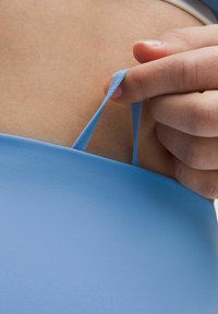 Gaiši zilas elastīgas auduma, ar gludu tekstūru; roka tur plānu siksnu tuvu ķermenim, izceļot apģērba dizainu un piegriezumu.