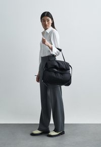 Sac à épaule noir rembourré avec des sangles réglables, forme arrondie et zip. Porté par une personne en pantalon gris et chemise blanche.