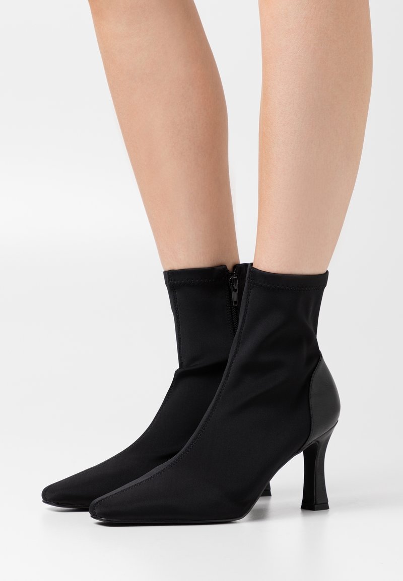 Schwarze Knöchelstiefel aus dehnbarem Stoff mit spitz zulaufender Zehenpartie, Reißverschluss und gebogenem Absatz, mit glatter Textur und minimalen Details.