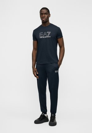 T-shirt bleu marine à manches courtes avec un logo graphique blanc. Pantalon de jogging assorti bleu marine avec un logo sur la cuisse gauche. Baskets noires.