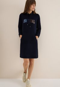 Mörkblå hoodieklänning med stor textgrafik "NATURAL", framfickor, dragsko i huvan och ribbade ärmslut och nederkant. Bärs med beiga sneakers.