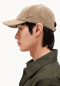 Beige Cordhosen-Baseballmütze mit gebogenem Schirm, sechs Panelen und verstellbarem Riemen; getragen von einer Person vor einem weißen Hintergrund.