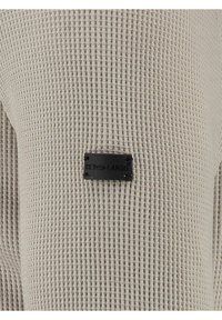 Beige gebreide trui met textuur, voorzien van een zwart rechthoekig merklabel met "KEY LARGO" reliëf, bevestigd op de borst.