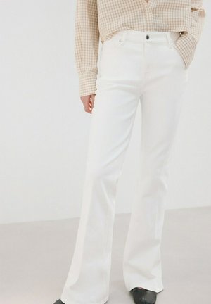 Persona con pantalones acampanados blancos de talle alto, camisa de manga larga a cuadros beige metida por dentro y zapatos negros, de pie contra un fondo blanco.