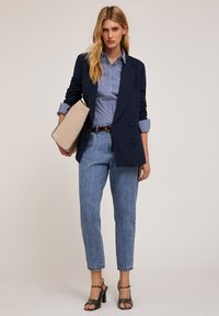 Granatowa marynarka z podwójnymi guzikami, jasnoniebieska w paski koszula, jasnoniebieskie dżinsy i czarne sandały na obcasie. Beżowa torebka z czarnym wykończeniem.