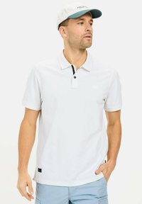 Hvid polo-shirt med en sort knapstolpe, korte ærmer og et lille logo på brystet; parret med lyseblå shorts og en kasket.