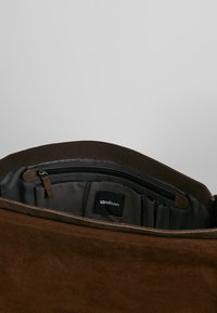 Hnědá suede taška s hladkou vnitřní podšívkou, obsahující zipovou kapsu a několik přihrádek, s černou etiketou uvnitř.