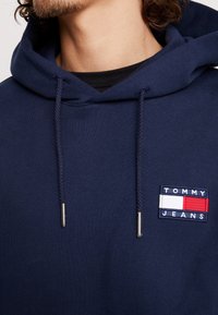 Sudadera con capucha azul marino con cordones, material de algodón. Presenta un logo rectangular bordado en rojo, blanco y azul en el pecho.