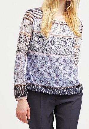Kvinde iført en mønstret bluse med lange ærmer med blomster- og geometriske designs og sorte bukser, stående mod en ensfarvet baggrund.
