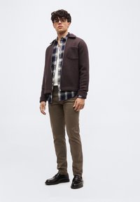 Νέος άνδρας με γυαλιά, φοράει ένα σκούρο καφέ μπουφάν με φερμουάρ πάνω από ένα μπλε καρό πουκάμισο, καφέ παντελόνι και μαύρα παπούτσια, στέκεται μπροστά σε ανοιχτόφωρο φόντο.