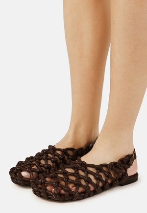 Sötétbarna bőr szőtt szandált viselő lábak, bokapántokkal, lábujjak láthatók a nyitott rácsminta között.