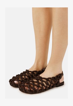 Pieds portant des sandales en cuir tressé brun foncé avec des brides à la cheville, laissant voir les orteils à travers un design en treillis ouvert.