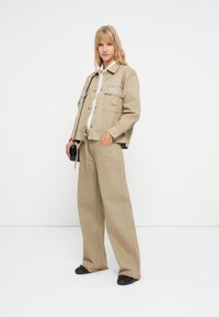 Beige denimjakke og bukser med vide ben med kontraststing, kombinert med en hvit skjorte og svarte sko, poserer mot en hvit bakgrunn.