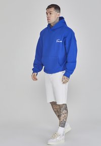 Hoodie in cotone blu con tasca frontale e logo ricamato bianco, abbinato a pantaloni corti di denim bianchi e sneakers bianche. Maniche corte, vestibilità comoda.