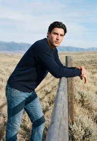 Homme en pull bleu marine et jean appuyé sur une clôture en bois dans une prairie sèche ouverte avec des montagnes en arrière-plan sous un ciel clair.
