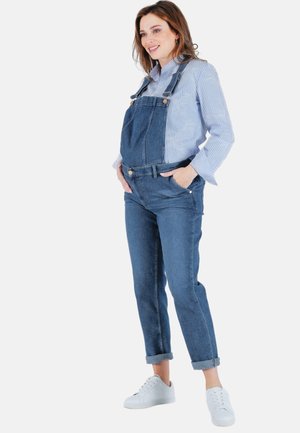 Salopette en denim bleu foncé avec des bretelles ajustables, une poche frontale, des jambes fuselées et des ourlets roulés, accompagnée d'une chemise rayée bleu clair et de baskets blanches.