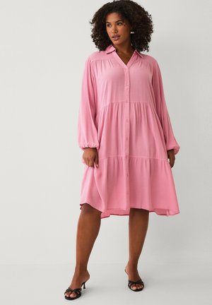 Kvinde iført en løs, langærmet, knappet pink midikjole med krave og lagdelt nederdel, kombineret med sorte sandaler med hæl.